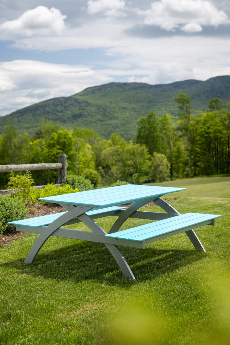 A light blue picnic table on green grass with lush mountains in the background at Plymouth Bay under a partly cloudy sky.