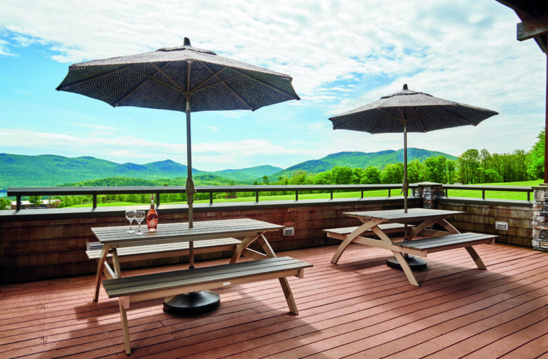 Two wooden picnic tables with umbrellas on a sunny terrace overlooking a scenic landscape of green mountains and Plymouth Bay under clear skies.