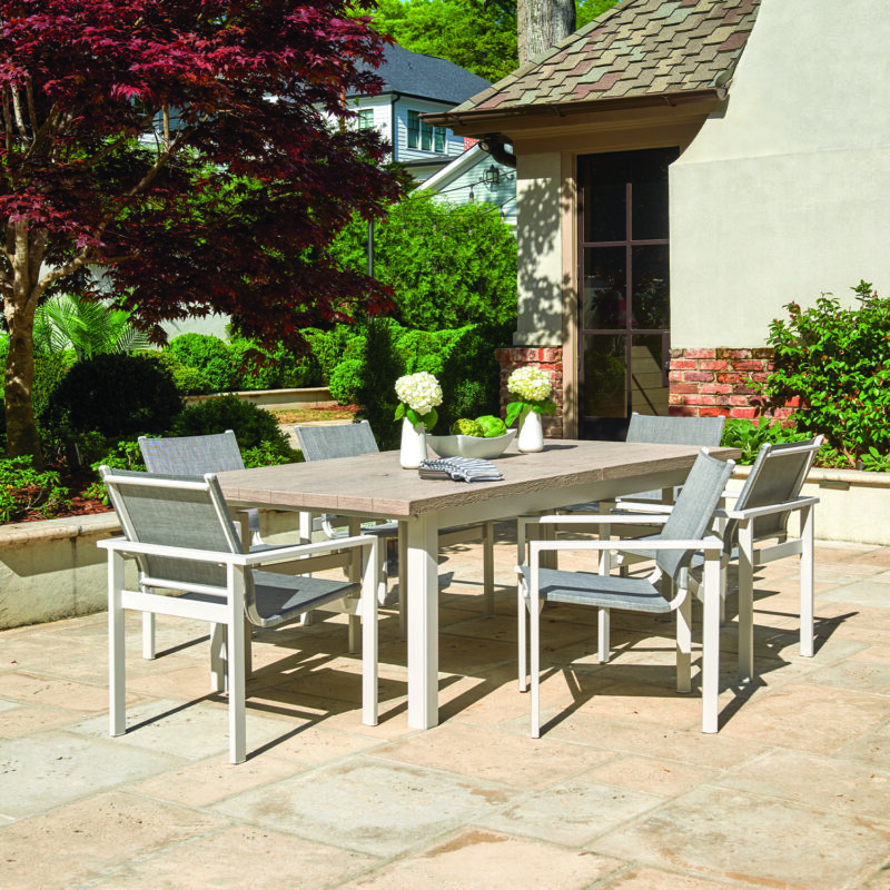 Outdoor dining area with a wooden table and six modern chairs on a patio surrounded by lush greenery and a red maple tree.