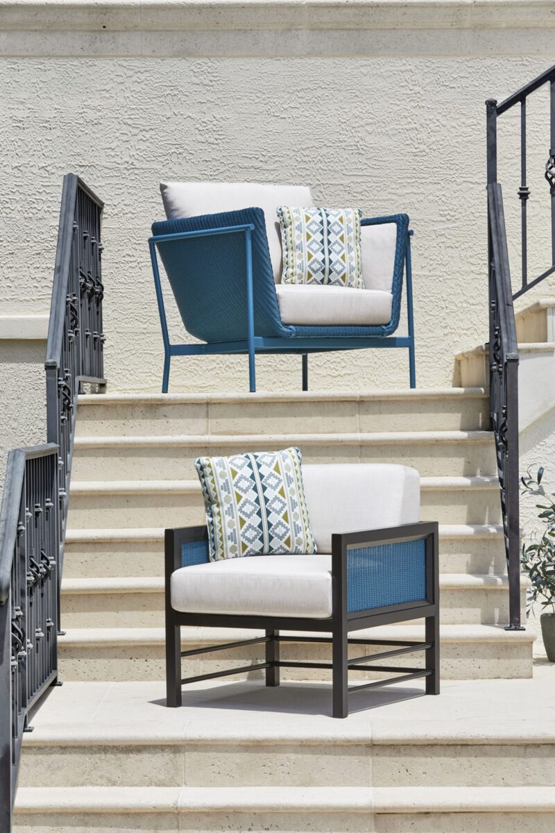 Two modern outdoor chairs with cushions, one blue and one black, arranged on concrete steps next to a black metal stair railing, under bright sunlight.