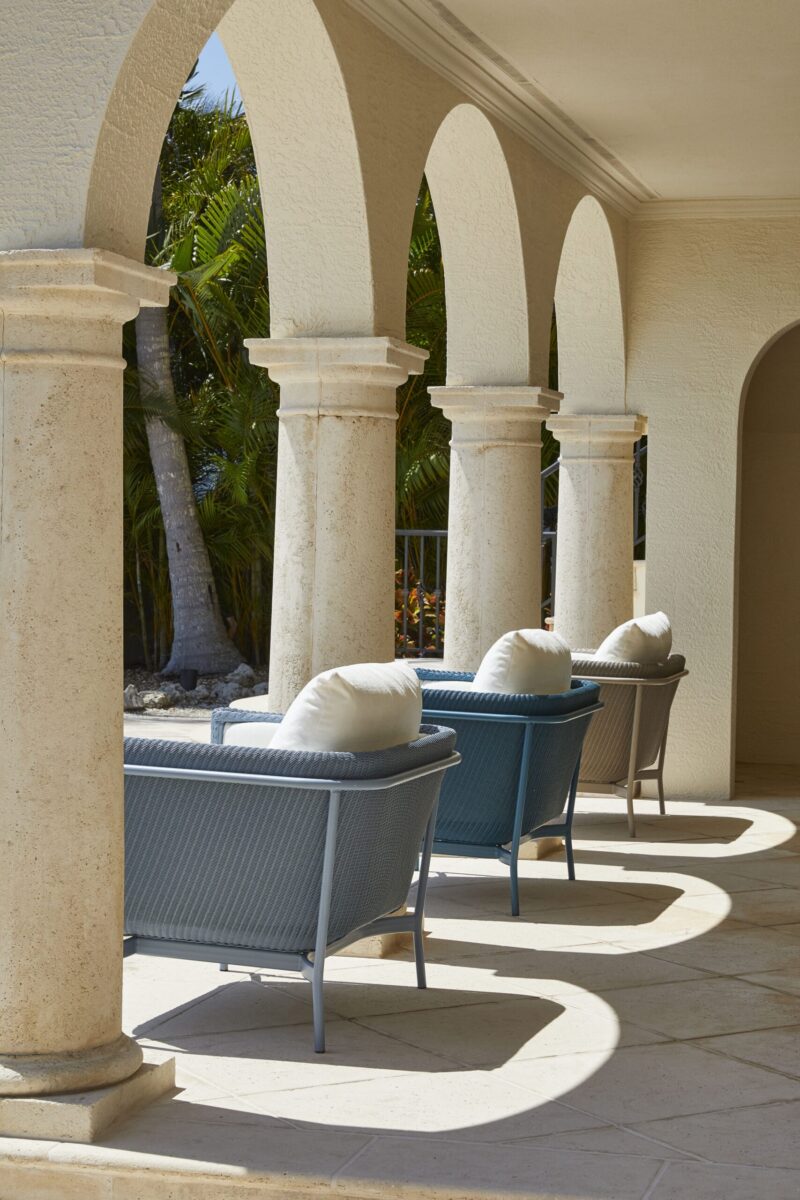 A sunlit patio featuring a series of archways with sturdy columns, lined with blue wicker chairs topped with white cushions. shadows create elegant patterns on the tiled floor.