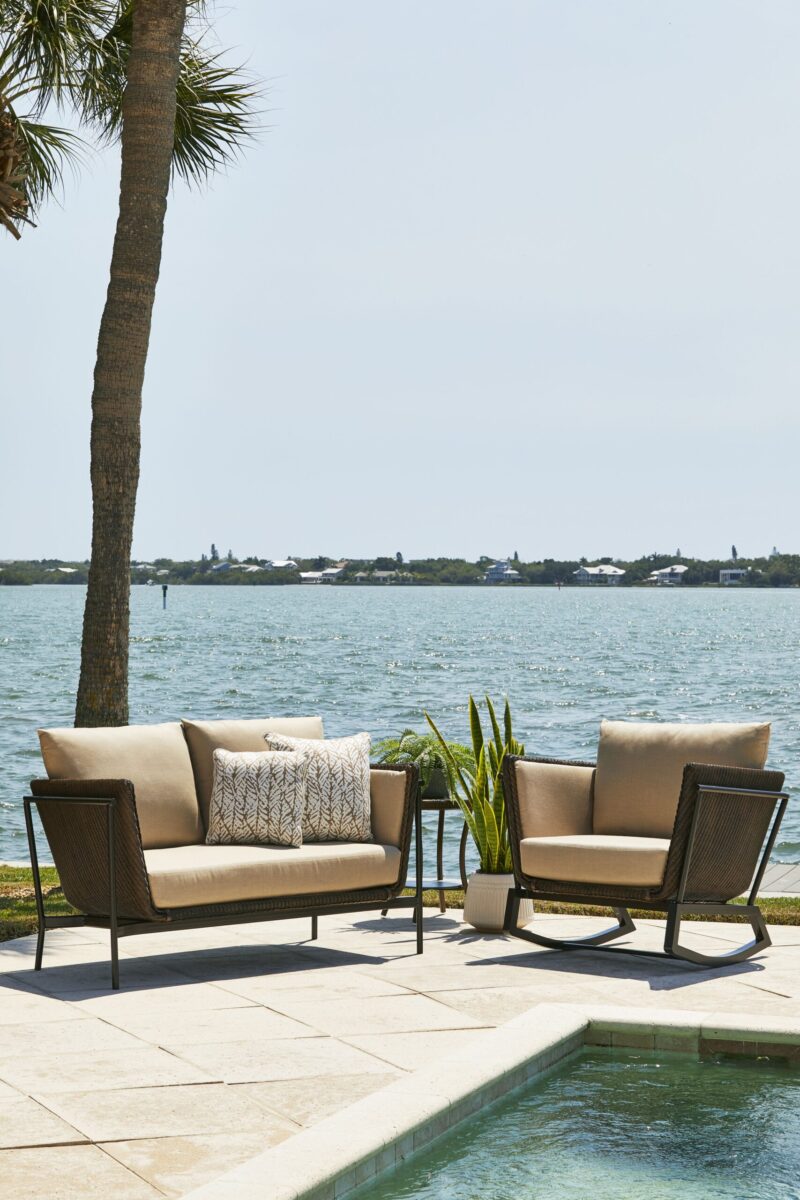 Outdoor patio setting by a pool with a couch and two chairs, overlooking a tranquil water body. a palm tree is to the left and a small green plant to the right, under sunny skies.