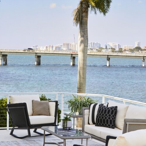 An outdoor seating area with modern furniture, overlooking a calm bay with a city skyline and a bridge in the background, framed by a palm tree.