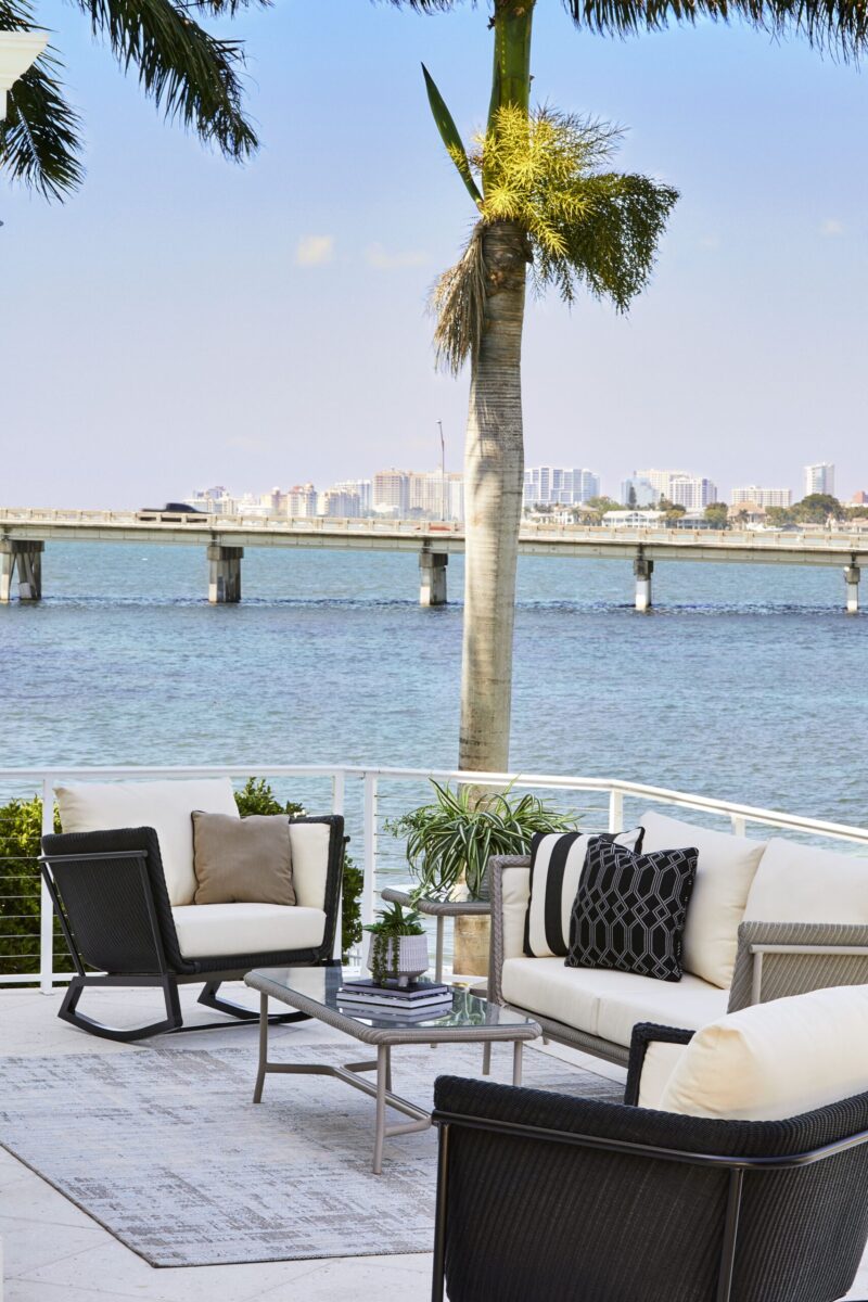 An outdoor seating area with modern furniture, overlooking a calm bay with a city skyline and a bridge in the background, framed by a palm tree.