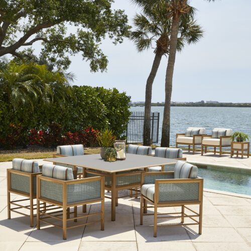 Outdoor lounge area with a sectional sofa and chairs around a coffee table, under palm trees, adjacent to a calm river. a swimming pool and more seating are visible in the background.