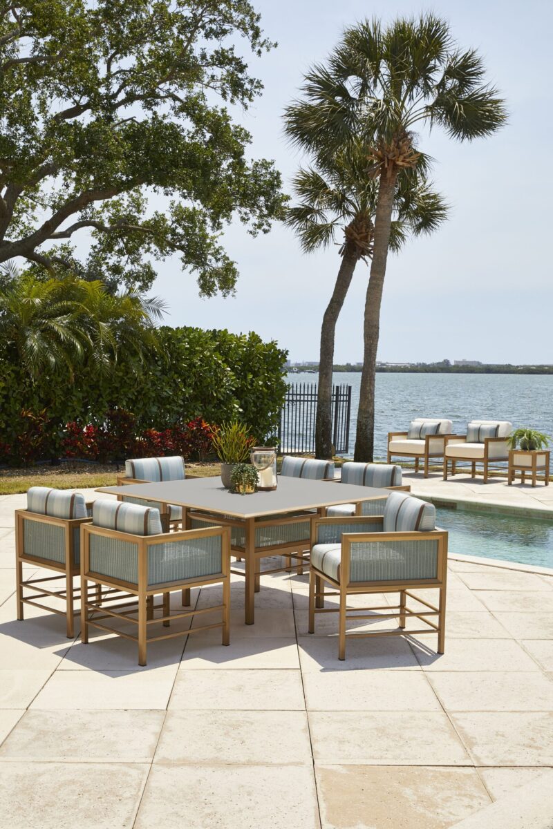 Outdoor lounge area with a sectional sofa and chairs around a coffee table, under palm trees, adjacent to a calm river. a swimming pool and more seating are visible in the background.
