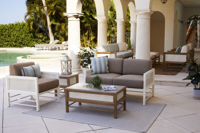 Luxurious outdoor patio furniture set featuring a sofa, chairs, and a coffee table arranged on a rug, with a pool in the background, surrounded by stylish columns and lush plants.