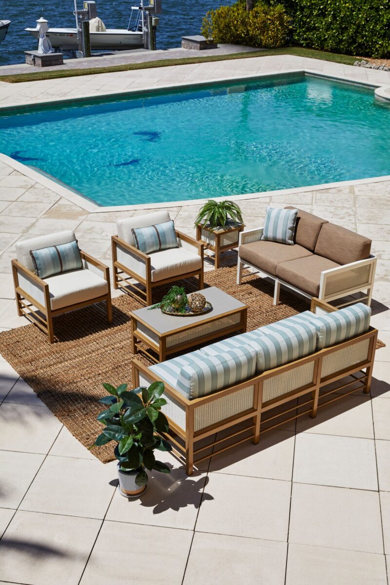 Outdoor patio furniture set by a swimming pool, featuring a sofa, chairs, and a coffee table on a textured rug, with a waterfront view and bright sunny day.