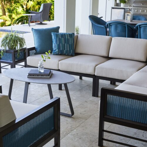 Elegant outdoor patio with beige sectional sofa and blue cushions, accompanied by matching armchairs and a coffee table with a plant, overlooking a sunny garden.
