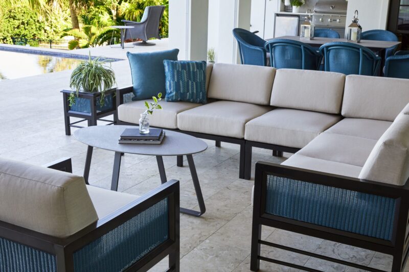 Elegant outdoor patio with beige sectional sofa and blue cushions, accompanied by matching armchairs and a coffee table with a plant, overlooking a sunny garden.