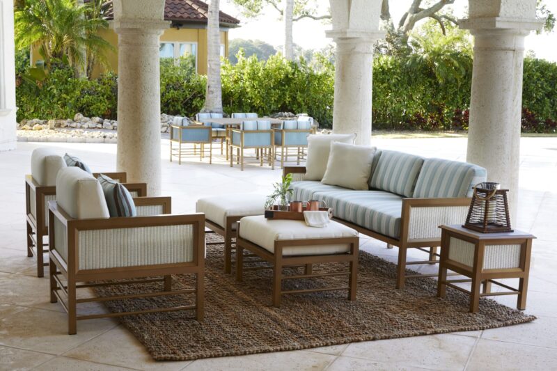 An elegant outdoor seating area featuring a sofa and multiple chairs with cushions, centered around a coffee table on a rug, under a pillared patio.