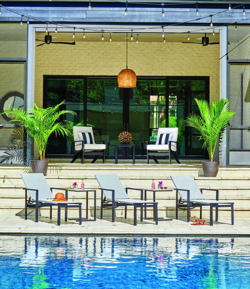 A luxurious poolside patio with comfortable lounge chairs, stylish outdoor seating, and decorative palm plants. a large hanging lamp and reflective windows on a building enhance the serene setting.