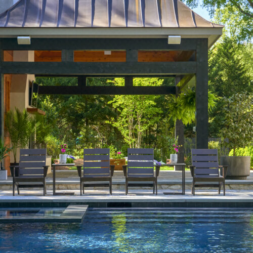 A serene backyard with a swimming pool and a shaded pavilion containing a dining set. the pool’s edge is lined with stylish chairs facing a lush garden and elegant residential structures.