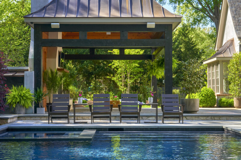 A serene backyard with a swimming pool and a shaded pavilion containing a dining set. the pool’s edge is lined with stylish chairs facing a lush garden and elegant residential structures.