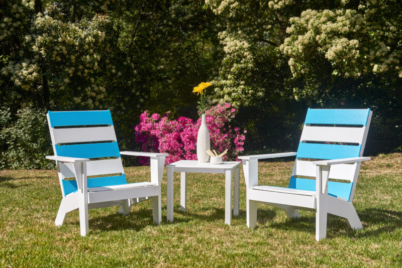 Two vibrant blue and white striped adirondack chairs facing each other with a small white table between them, set against blooming bushes in a sunny garden.