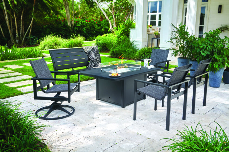 Modern outdoor dining area with a black table and chairs set on a stone patio, surrounded by a lush green garden and a white house exterior.