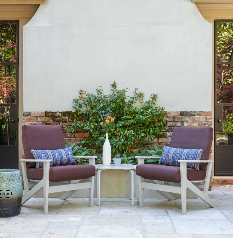Two outdoor chairs with cushions and a small table between them, featuring a vase, set against a garden wall with greenery and a retractable awning above.