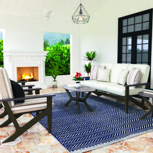 A cozy indoor patio with a beige couch, striped cushions, a geometric coffee table, a side chair, and a fireplace. the setting features tile flooring, large windows, and views of greenery outside.