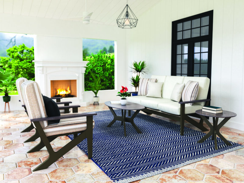 A cozy indoor patio with a beige couch, striped cushions, a geometric coffee table, a side chair, and a fireplace. the setting features tile flooring, large windows, and views of greenery outside.