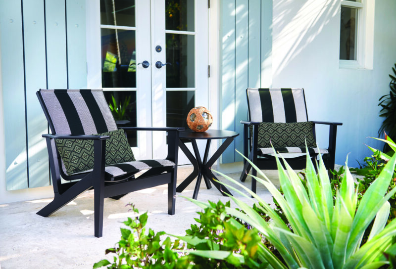 Two black and white patterned outdoor chairs and a small round table with a decorative vase, beside a light blue door and surrounded by lush green plants.