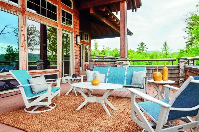 A cozy patio setting with modern white and blue furniture on a wooden deck, surrounded by large glass windows and lush greenery, under a clear sky.