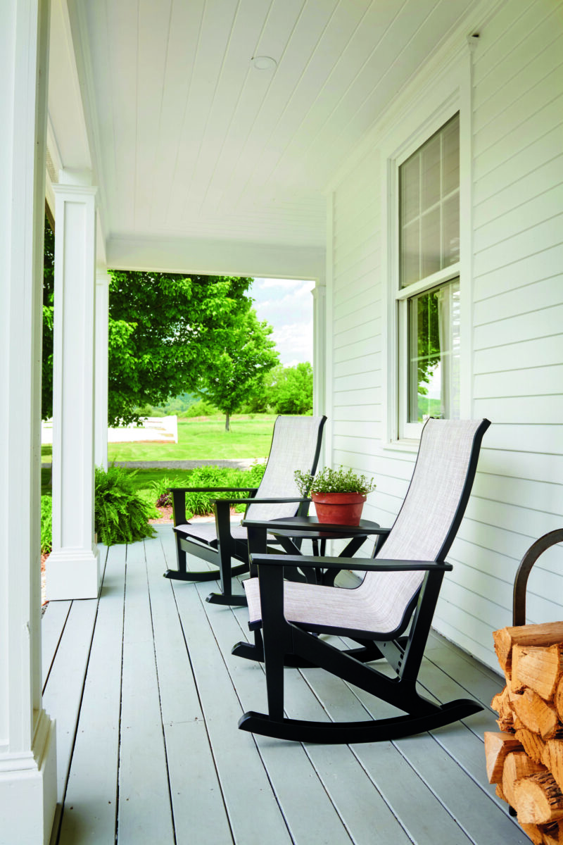 Two rocking chairs on a bright, white porch with a view of a lush green lawn and trees outside. a small wooden table with plants sits between the chairs.