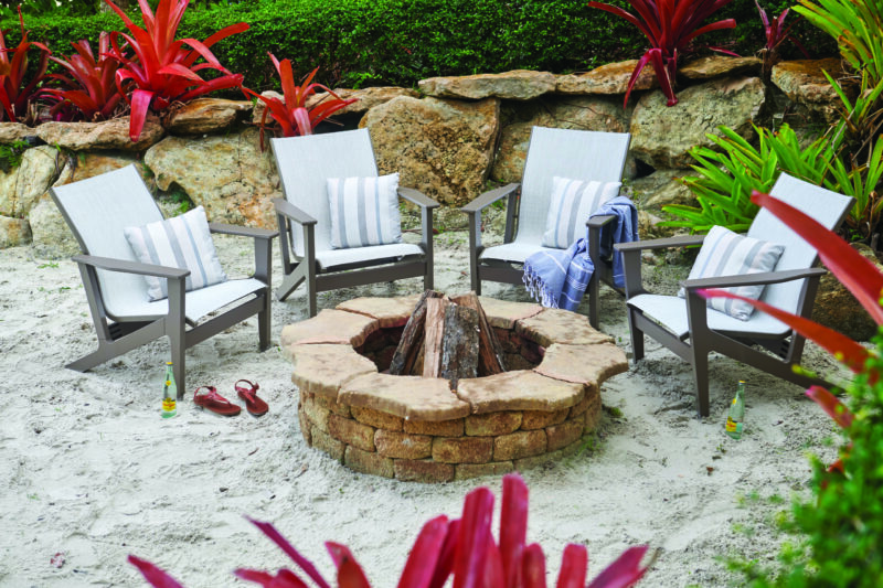 Four modern chairs around a stone fire pit in a sandy area with lush red plants and rocks in the background. comfortable outdoor setting for relaxation.