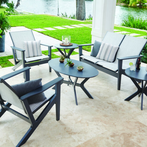 Modern outdoor patio furniture set by a lake, including chairs with cushions and two tables with decorative items, in a lush garden setting.