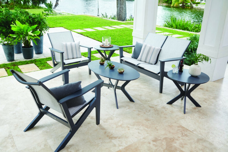 Modern outdoor patio furniture set by a lake, including chairs with cushions and two tables with decorative items, in a lush garden setting.