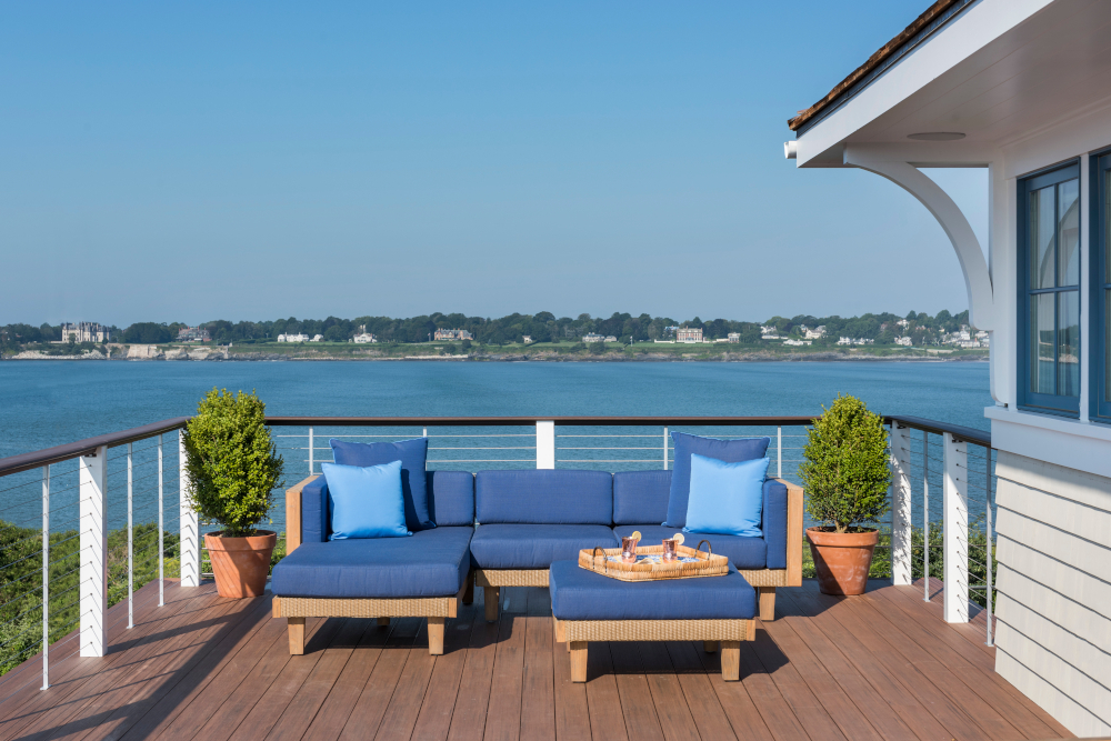Spacious waterfront deck featuring a cozy outdoor seating area with blue cushions, a small table, and a panoramic view of the calm Catalina sea and distant shoreline under a clear blue sky.