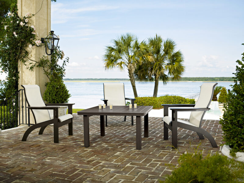 Outdoor patio with modern furniture overlooking a scenic lake, featuring a set of white cushioned chairs and a table set on a cobblestone floor, surrounded by greenery and a palm tree.