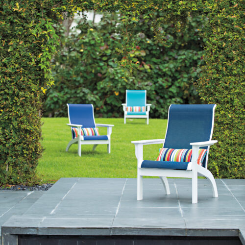 An outdoor setting featuring white chairs with colorful striped cushions by a pool, surrounded by lush green hedged walls.