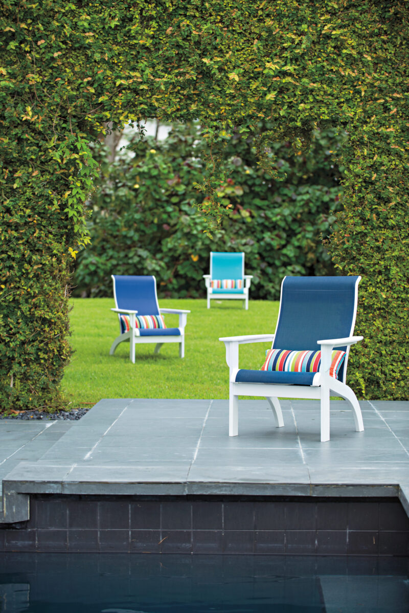 An outdoor setting featuring white chairs with colorful striped cushions by a pool, surrounded by lush green hedged walls.