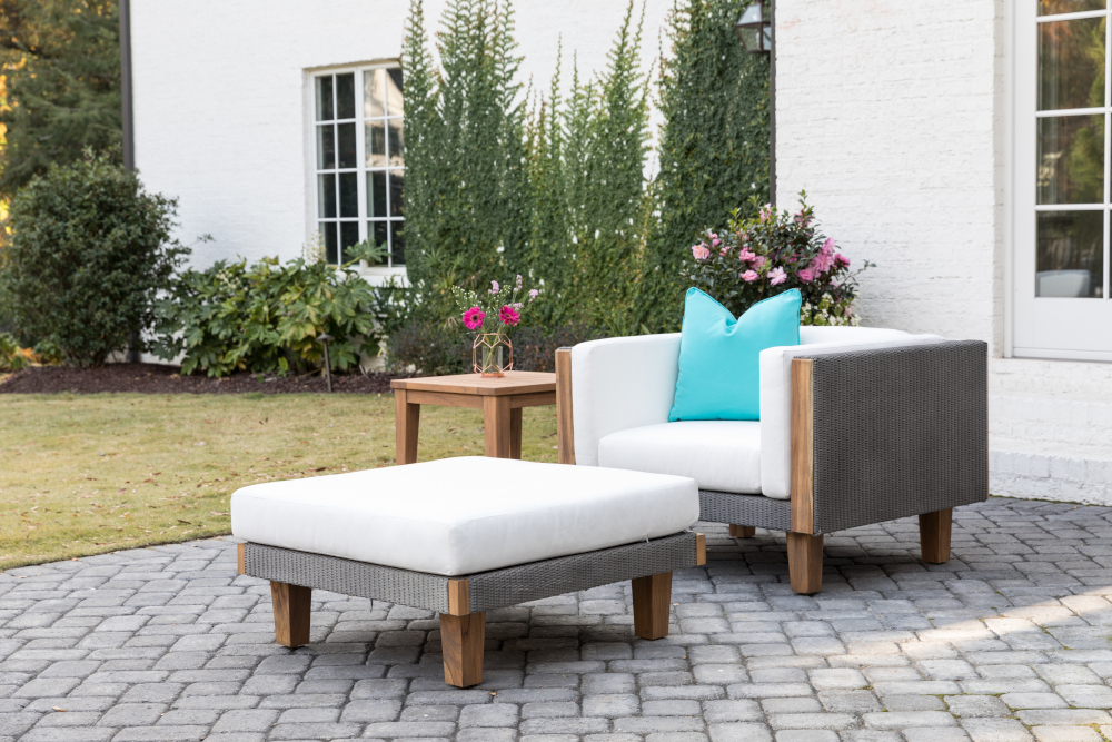 Two modern Catalina outdoor chairs with white cushions and a matching ottoman on a paved patio, near a plant-filled table, against a backdrop of a white house and tall green bushes.