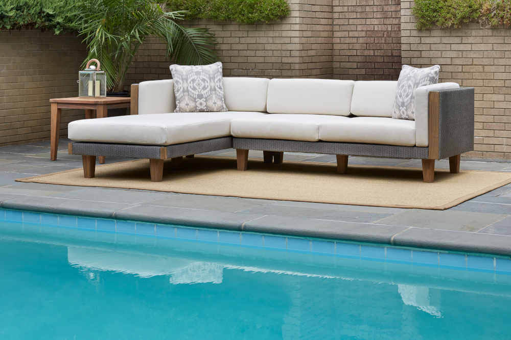 A modern outdoor sectional sofa with white Catalina cushions on a beige rug by a swimming pool, surrounded by a brick wall and complemented by a wooden side table.