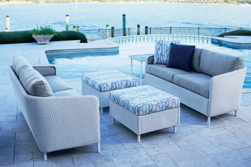 Outdoor patio furniture set by a pool, including a sofa, armchairs, and ottomans with gray woven frames and blue-gray cushions, beside a calm body of water.