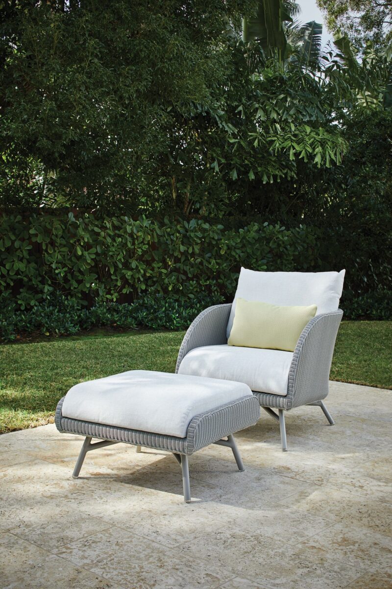 A modern outdoor lounge chair with a matching ottoman, set on a stone patio surrounded by lush greenery. the chair is gray with white cushions and a pale green pillow.
