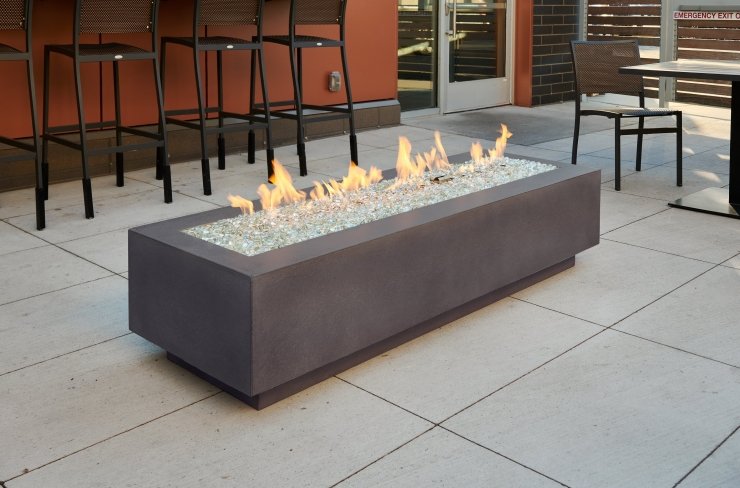 A modern outdoor rectangular fire pit with a blazing flame, surrounded by glass pebbles, situated on a patio area with seating in the background.