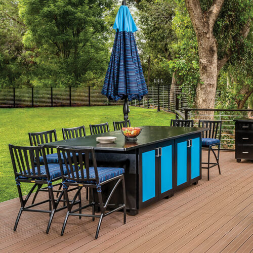Outdoor dining setup on a wooden deck featuring a Gensun Amari black table with blue accents, matching chairs, and a large blue umbrella, surrounded by lush greenery and a wooden fence.