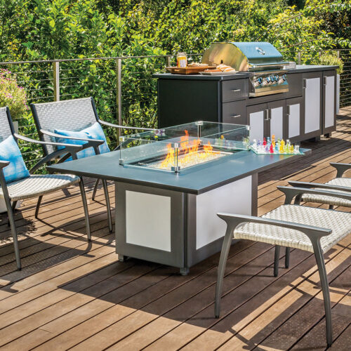 Modern outdoor patio featuring a Gensun Amari dining set, including a table with a built-in fire pit, surrounded by chairs and a cooking grill, set on a wooden deck overlooking green foliage.
