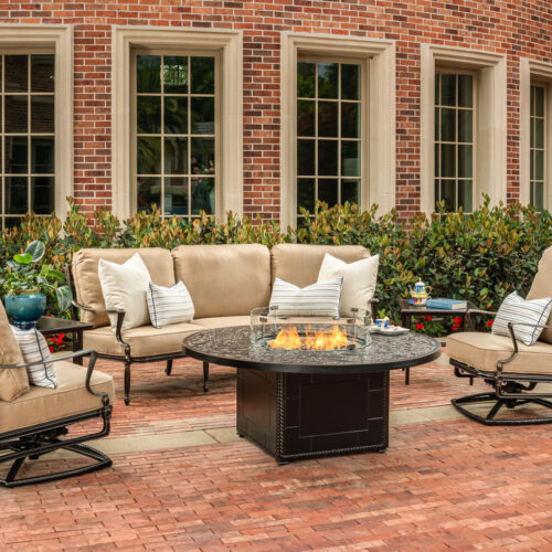 An outdoor seating area with a Gensun Bel Air fire pit table, surrounded by a sofa and two swivel chairs on a brick patio, adjacent to a red brick building with large windows.