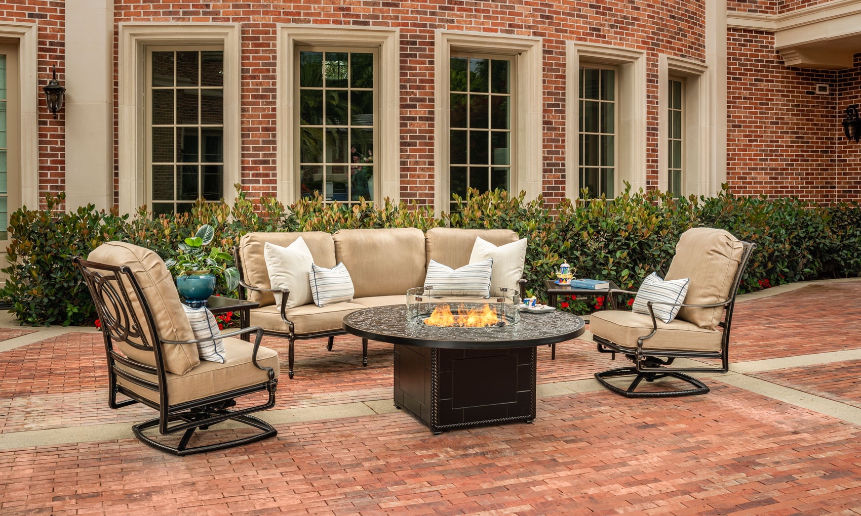 An outdoor seating area with a Gensun Bel Air fire pit table, surrounded by a sofa and two swivel chairs on a brick patio, adjacent to a red brick building with large windows.