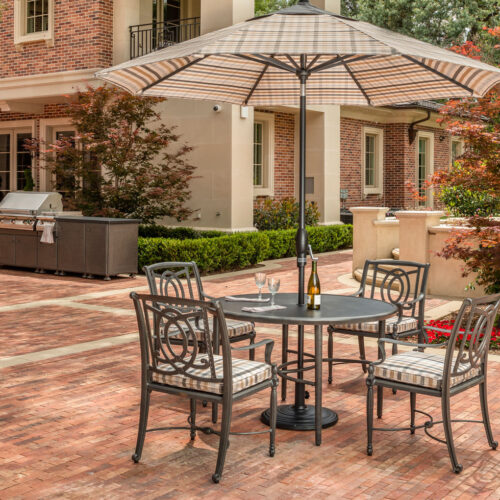Patio area with a Gensun Bel Air dining set under an umbrella next to a brick house, surrounded by trees and a barbecue grill on the side.