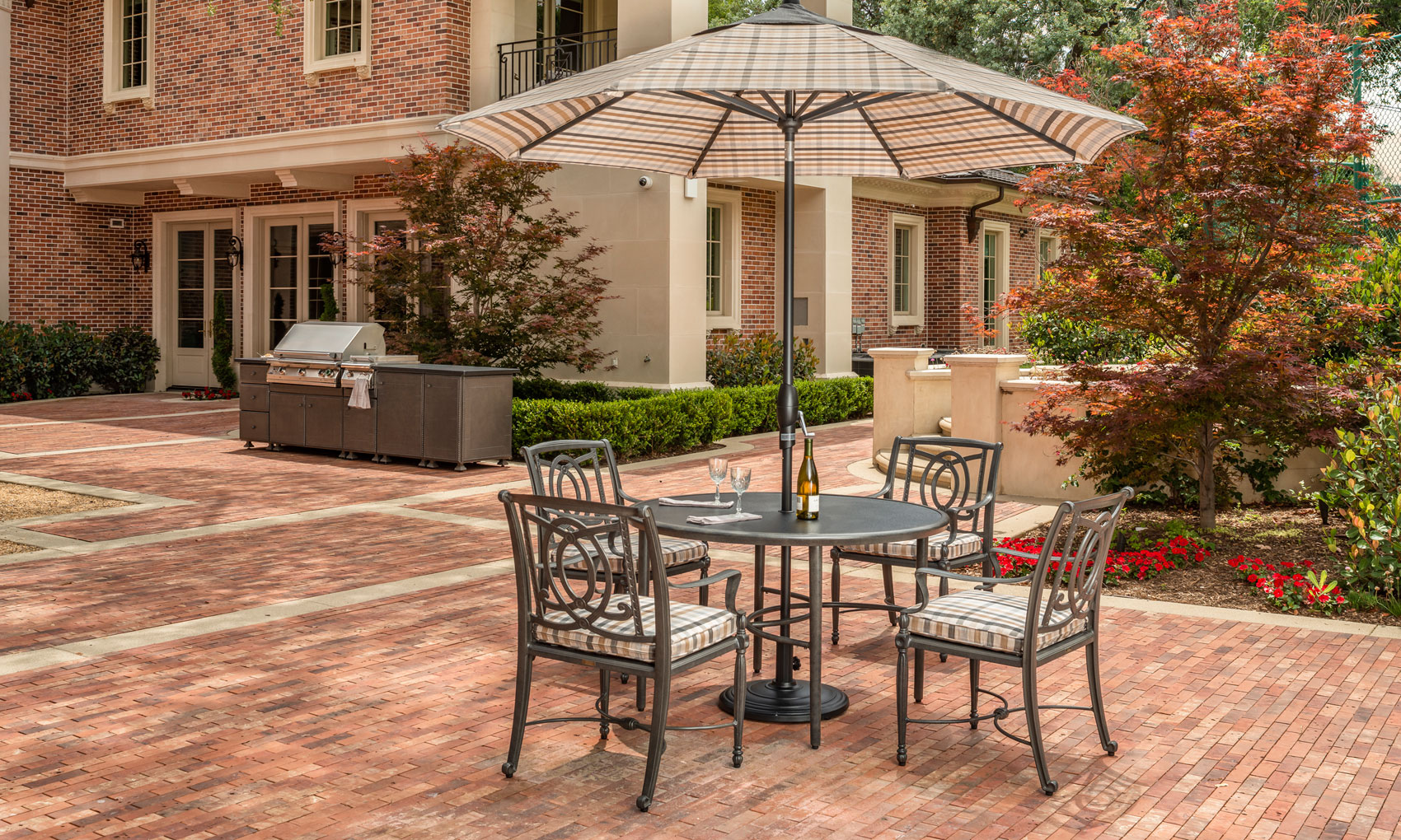 Patio area with a Gensun Bel Air dining set under an umbrella next to a brick house, surrounded by trees and a barbecue grill on the side.