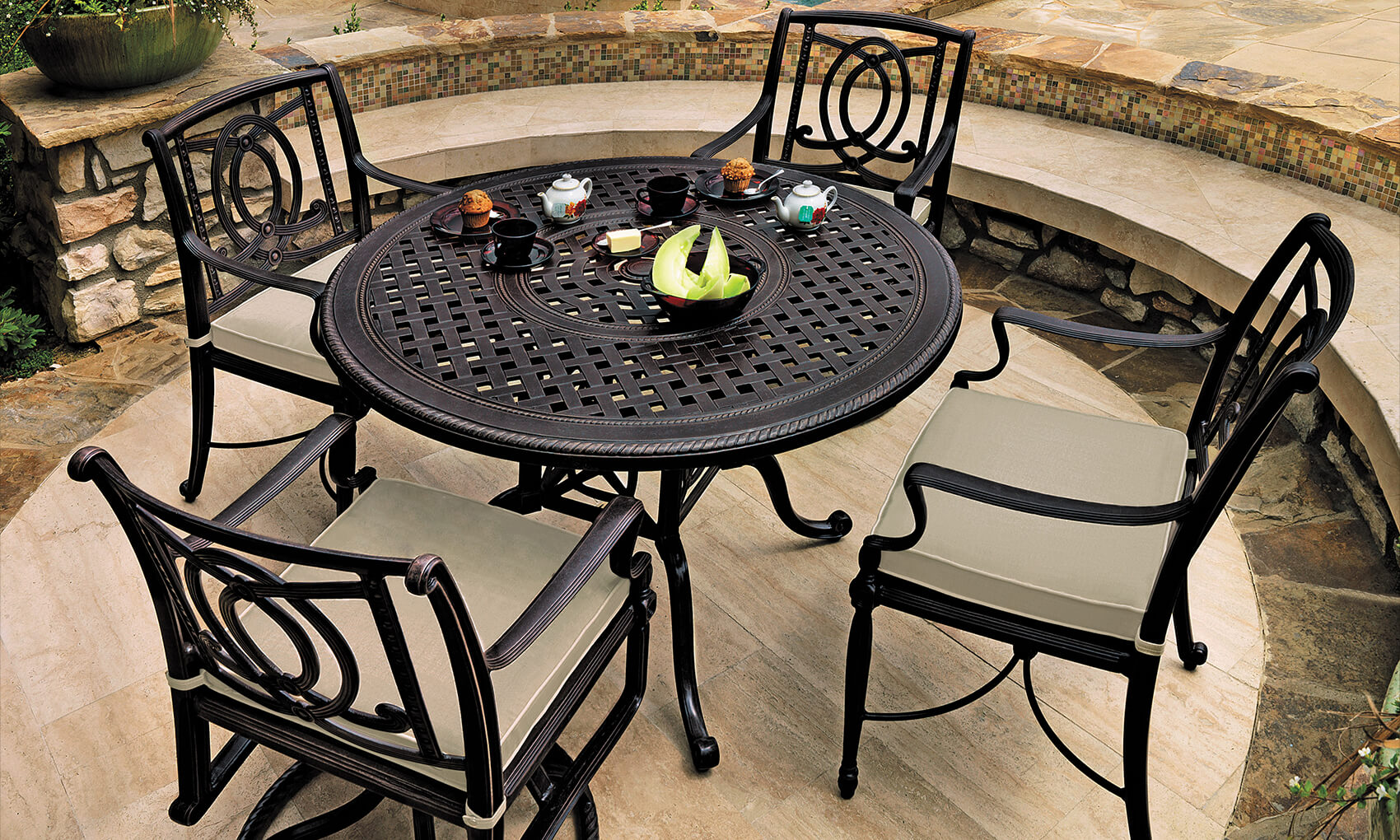 An outdoor dining set from the Gensun Bel Air collection, consisting of a round, ornate metal table and four matching chairs on a stone patio, with cups, plates, and a centerpiece of