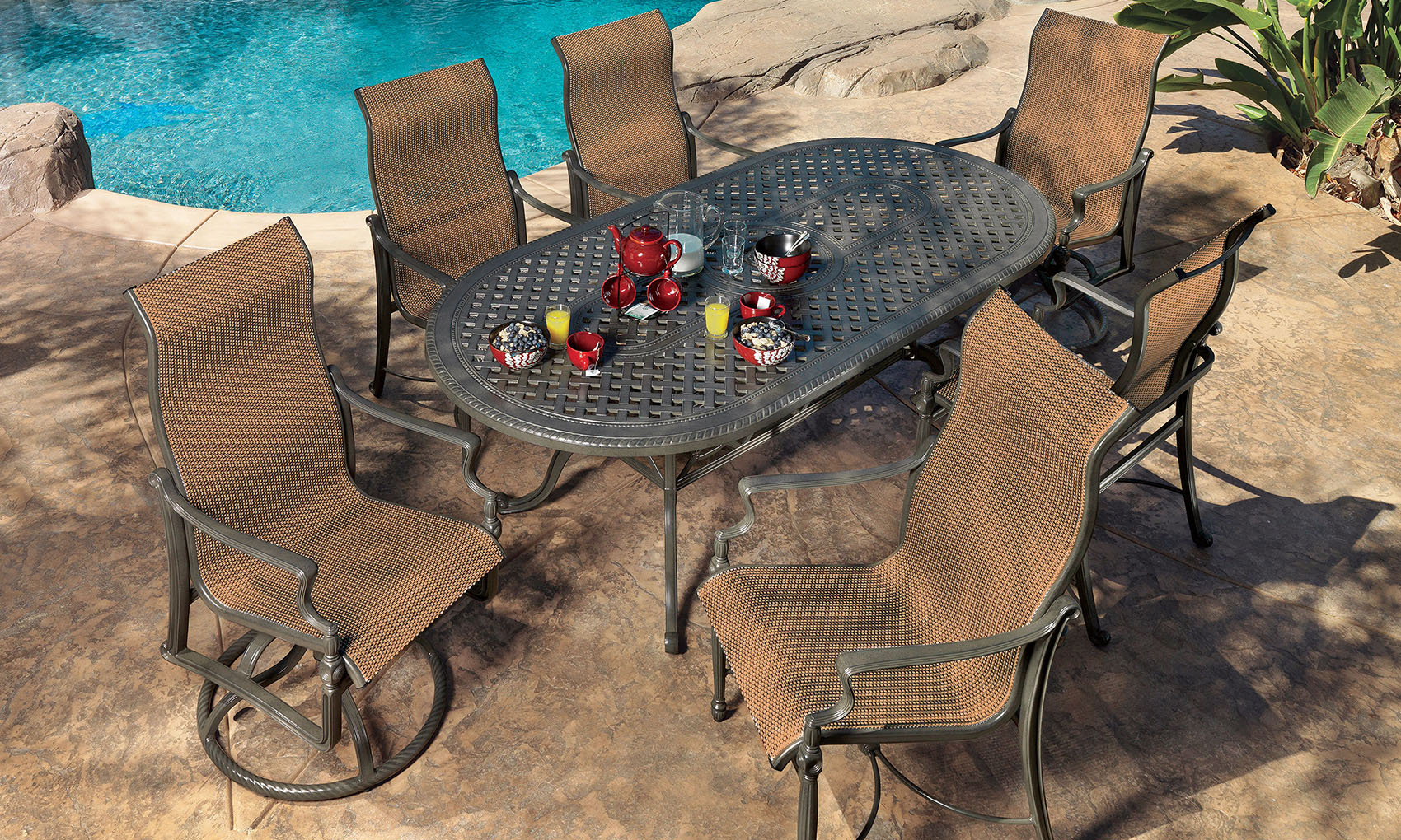 Outdoor dining area with a Gensun Bel Air metal table set for six, featuring wicker chairs near a pool surrounded by rocks and lush greenery.