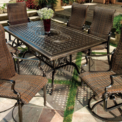 Outdoor patio setting featuring a Gensun Bel Air wrought iron and wicker table surrounded by four matching chairs on a stone pavement, adorned with a small flower arrangement in the center.