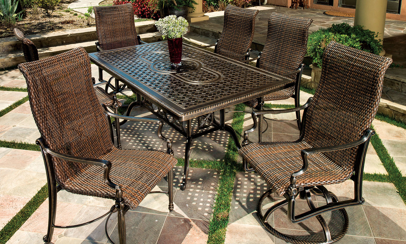 Outdoor patio setting featuring a Gensun Bel Air wrought iron and wicker table surrounded by four matching chairs on a stone pavement, adorned with a small flower arrangement in the center.