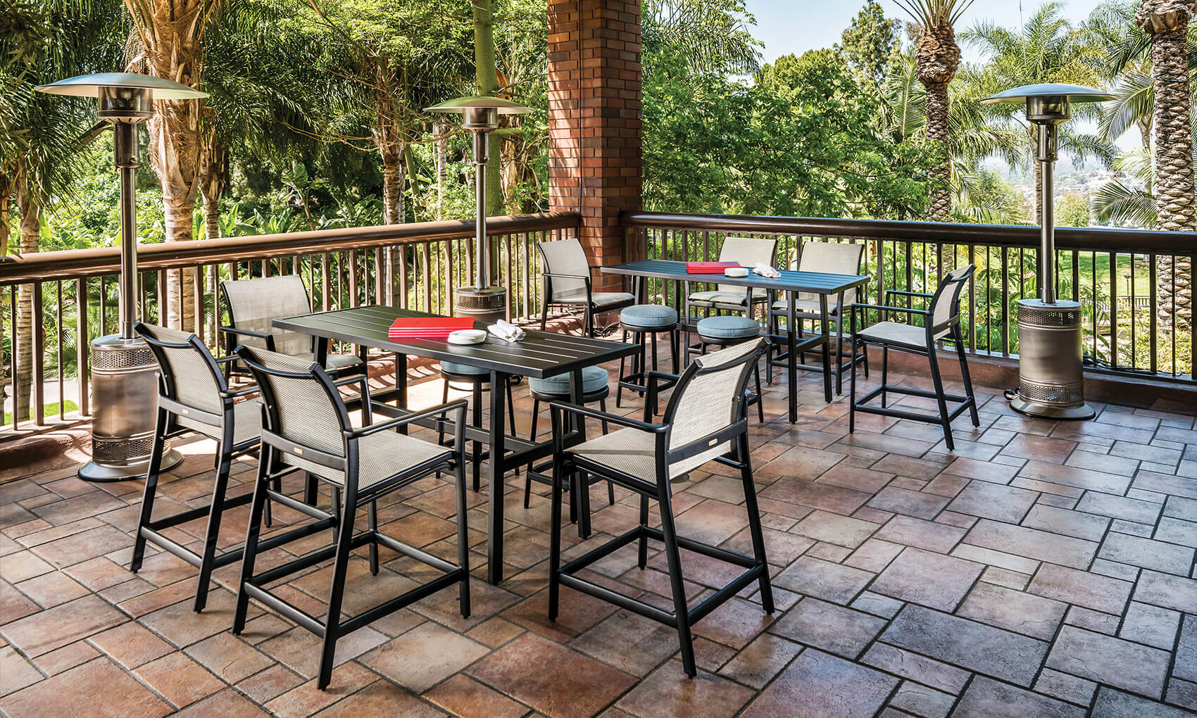 Spacious outdoor terrace with a Gensun Echelon dining area featuring multiple tables and chairs on a terracotta tiled floor, surrounded by lush greenery and equipped with patio heaters.