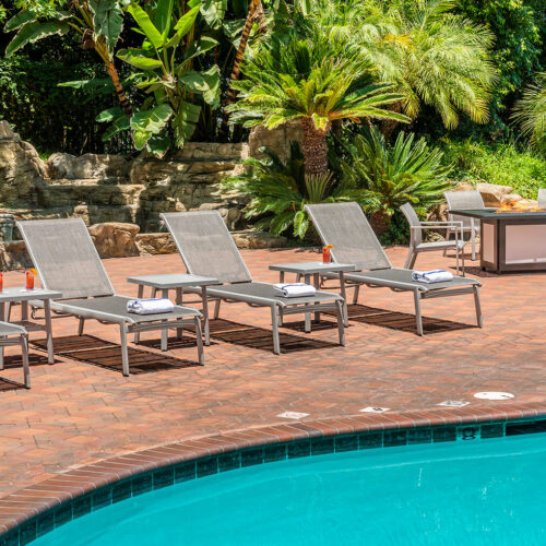 Four Gensun Echelon lounge chairs by a swimming pool, with towels and a scenic, lush garden background. A small round table and additional patio seating are visible near the pool.